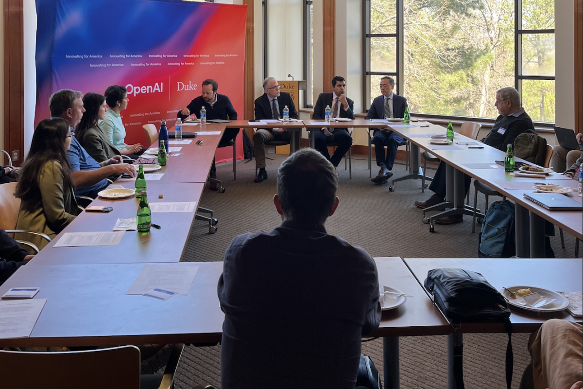 A group of people sits around a U-shaped conference table in a well-lit room with large windows. At the front, four panelists are engaged in a discussion, with a backdrop displaying the logos of OpenAI and Duke University. Bottled water and papers are on the tables, and attendees are listening attentively. The atmosphere suggests an academic or policy-focused roundtable event.