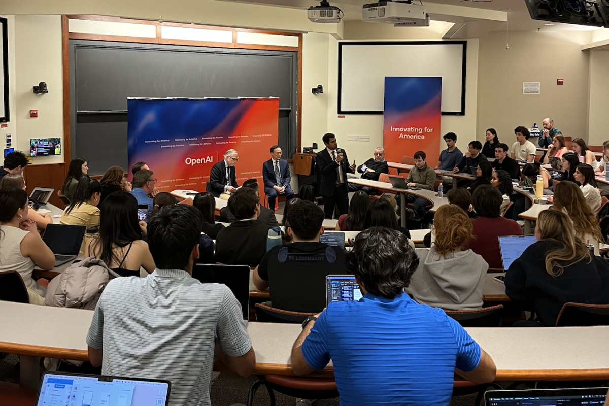 A large audience of students and attendees sits in a lecture hall, facing a panel discussion in front of two OpenAI-branded banners that read "Innovating for America." Four panelists are seated at the front, with one person standing and speaking with a microphone. Many audience members are using laptops, and the atmosphere is engaged and attentive, suggesting a university event focused on AI and public policy.