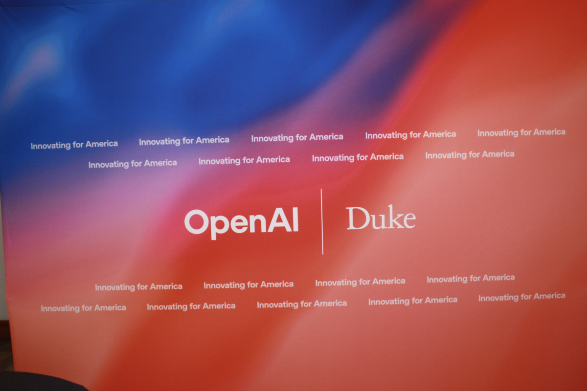   A vibrant red and blue gradient backdrop featuring repeated text that reads “Innovating for America.” Centered on the backdrop are the logos of OpenAI and Duke, separated by a vertical line, indicating a partnership or collaborative event between the two institutions.